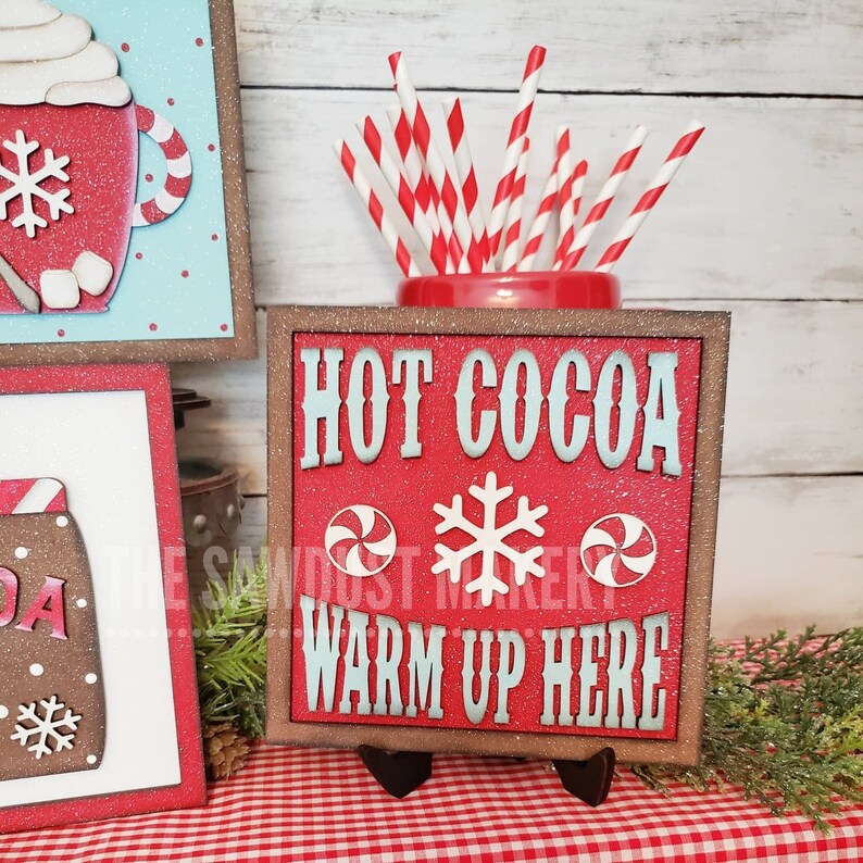 May include: Red wooden sign with white glitter and the text "HOT COCOA WARM UP HERE" in blue lettering. The sign has a white snowflake and two red and white peppermint candies on it. The sign is on a small black easel.