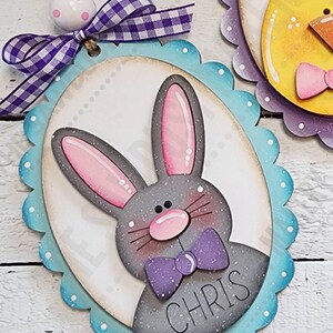 May include: A gray bunny with pink ears and a purple bow tie is on a blue and white oval-shaped wooden sign with the name "CHRIS" written on it. The sign has a scalloped edge and is decorated with white dots.