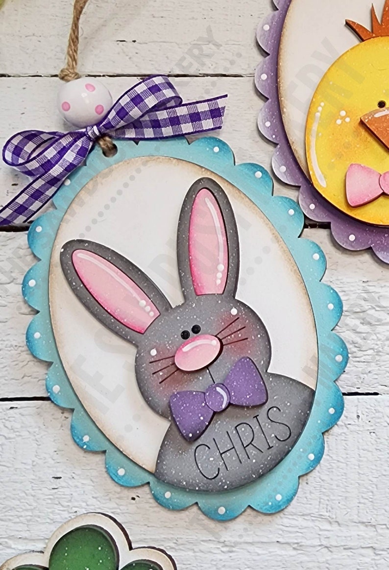 May include: A gray bunny with pink ears and a purple bow tie is on a blue and white oval-shaped wooden sign with the name "CHRIS" written on it. The sign has a scalloped edge and is decorated with white dots.