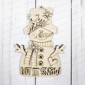 May include: A wooden snowman ornament with a hat, scarf, and buttons. The snowman has a carrot nose and a smiling face. The words "LET IT SNOW" are at the bottom. Includes snowflakes and a heart.