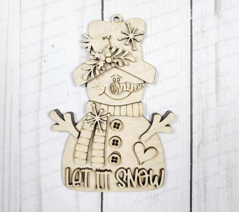 May include: A wooden snowman ornament with a hat, scarf, and buttons. The snowman has a carrot nose and a smiling face. The words "LET IT SNOW" are at the bottom. Includes snowflakes and a heart.
