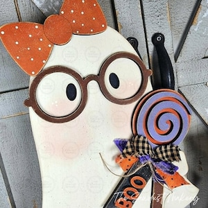 May include: A white ghost cutout decoration featuring a large orange bow with white polka dots, brown round glasses, and a purple and orange spiral lollipop. The ghost holds a sign with the orange text BOO and a decorative plaid fabric bow.