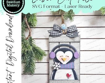 May include: A laser cut file for a penguin Christmas ornament. The ornament features a penguin wearing a scarf and earmuffs, with the words "seasons greetings" on the sign. The file is in SVG format and is laser ready.