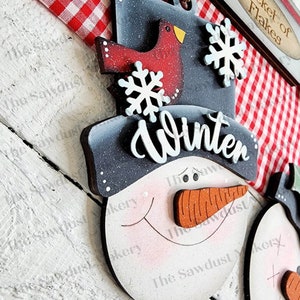 May include: Wooden snowman ornament with a black hat that says "Winter" in white lettering. The snowman has a red nose and a smiling face. There is a red bird on the hat and snowflakes on the hat and the snowman's face.