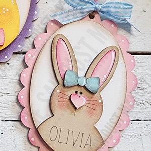 May include: A pink and white oval wooden Easter ornament with a brown bunny face, blue bow, and the name "Olivia" written on it.