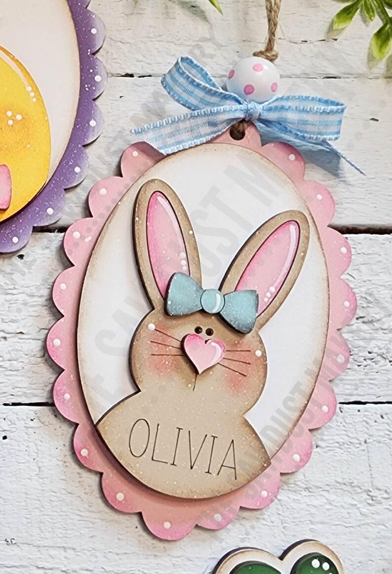 May include: A pink and white oval wooden Easter ornament with a brown bunny face, blue bow, and the name "Olivia" written on it.
