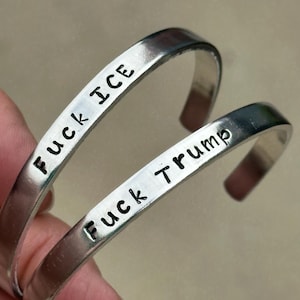 May include: Two silver-colored metal bracelets. One bracelet is stamped with the words "Fuck ICE", and the other is stamped with "Fuck Trump". The bracelets are held by a hand.