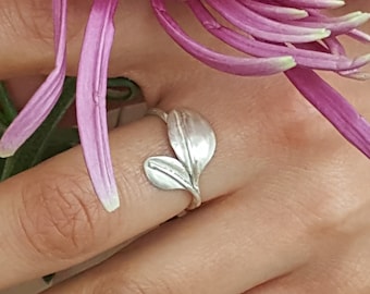 May include: A silver ring with a leaf design. The ring is on a finger and the background is a pink flower.