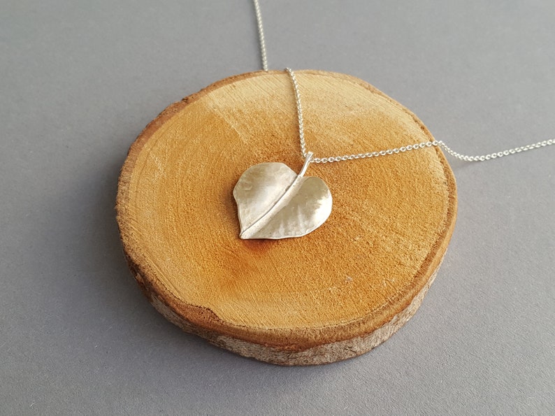 May include: A silver necklace with a hammered heart-shaped leaf pendant. The necklace is displayed on a wooden surface.