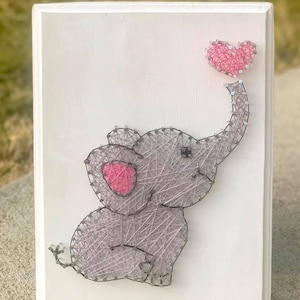 May include: A gray string art elephant holding a pink heart. The elephant is sitting on a white wooden background.