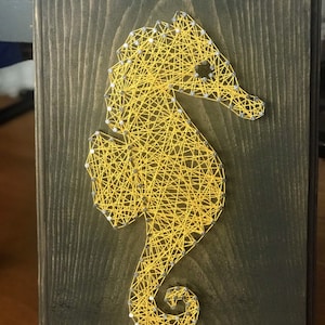 May include: Yellow string art of a seahorse on a dark wood background.