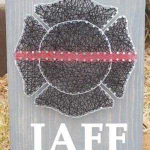 May include: A gray wooden sign with a black string art design of a firefighter's helmet. The helmet is outlined in silver nails and has a red string across the middle. The words "IAFF" are printed in white below the helmet.