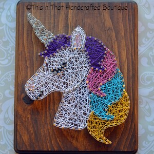 May include: String art of a unicorn's head with a white horn, purple, pink, blue, and yellow mane on a brown wooden background. The text "This n That Handcrafted Boutique" is in the top right corner.