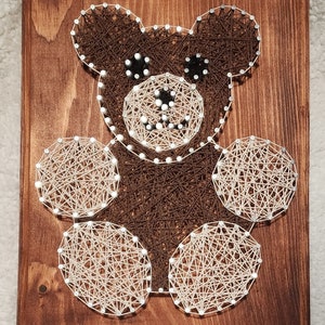 May include: A brown and white string art piece depicting a teddy bear with a smiling face. The bear is made of string wrapped around nails hammered into a wooden board.