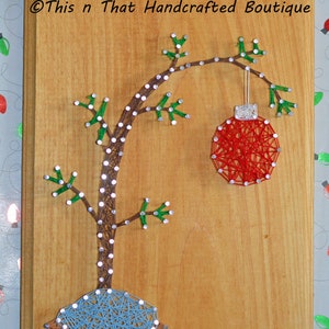 May include: Wooden plaque featuring a string art design of a Christmas tree with a red ornament. The tree trunk is brown, the branches are green, and the ornament is red. The base of the tree is blue. The text "This n That Handcrafted Boutique" is at the top.