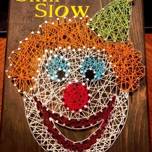 May include: A string art piece depicting a clown's face with a red nose, blue eyes, and a green hat. The background is a dark brown wood. The text "Slim Slow" is written in yellow letters at the top of the piece.