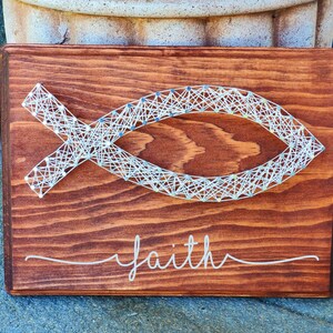 May include: A wooden sign with a white string art design of a fish with the word "faith" written below it.