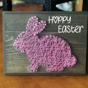 May include: Pink string art of a bunny rabbit on a dark brown wooden board with the text "Happy Easter" in white.