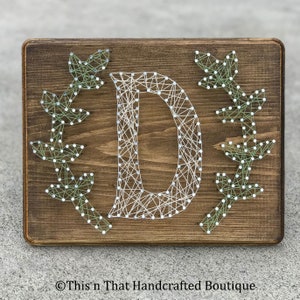 May include: A wooden sign with the letter D created with string art. The letter is white and the background is brown. There are green leafy branches on either side of the letter.