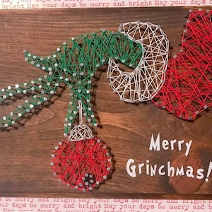 May include: A string art piece depicting the Grinch holding a red ornament. The background is a brown wooden board with the text "Merry Grinchmas!" written in white.