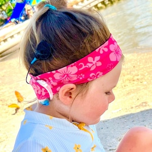以下が含まれることがあります： ピンクの花柄のヘアバンドと、黄色の花のアクセントが付いた水色と白のストライプの水着を着た幼い子供。 子供は補聴器を装着しています。