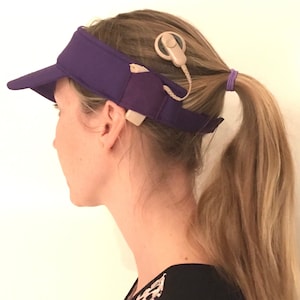 Könnte beinhalten: Ein lila Visier mit einem beigefarbenen Cochlea-Implantat. Das Implantat ist über ein Kabel mit einem Gerät am Kopf verbunden. Die Person trägt ein Oberteil mit schwarz-weißem Muster.