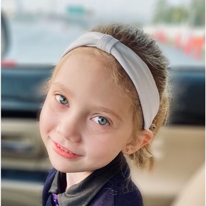 May include: A young person with blonde hair and blue eyes wearing a light gray knotted headband. The person is also wearing a dark purple collared shirt. The background is blurred, suggesting an outdoor setting.