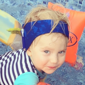 以下が含まれることがあります： 青と白のストライプの水着と、模様入りの青いヘアバンドをつけた子供。オレンジ色のアームフロートを使ってプールに入っています。