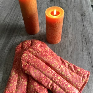 May include: Two lit orange pillar candles with a textured surface positioned behind a pair of quilted oven mitts featuring a repeating pattern of gold and orange circles on a pink background with stitched vertical channels for flexibility.