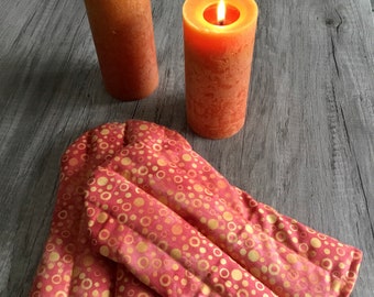 Könnte beinhalten: Zwei brennende orangefarbene Stumpenkerzen mit strukturierter Oberfläche hinter einem Paar gesteppter Ofenhandschuhe mit einem sich wiederholenden Muster aus goldenen und orangefarbenen Kreisen auf rosa Grund und vertikalen Steppnähten.