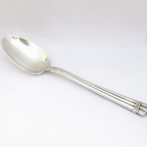 May include: A silver spoon with a rounded bowl and a long, slender handle. The handle features three parallel lines running down its length, with a decorative design at the end. The spoon is set against a white background.