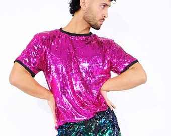 Könnte beinhalten: Eine Person trägt ein pinkfarbenes, sequinenbesetztes T-Shirt mit kurzen Ärmeln und eine grüne und blaue, sequinenbesetzte Hose. Die Person steht mit der rechten Hand auf der Hüfte und der linken Hand an der Seite. Sie trägt schwarze Turnschuhe.