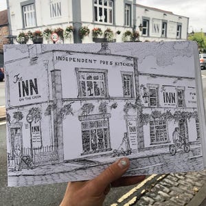 Op de afbeelding: Een zwart-wit pen- en inkttekening van een pub genaamd "The Inn on the Green". De tekening wordt voor de echte pub gehouden. De pub heeft hangende bloembakken en de tekst "Independent Pub & Kitchen".