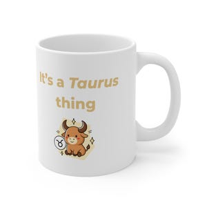 Puede incluir: Taza de cerámica blanca con el texto "It's a Taurus thing" en letras doradas. La taza presenta una ilustración de un toro de dibujos animados con el símbolo del zodiaco Tauro. El diseño incluye acentos de estrellas doradas.