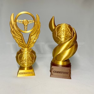 May include: Two gold-colored trophies. One features a steering wheel design with wings, a sphere, and the word "CHAMPION." The other has a twisted design with a sphere and the word "CHAMPION" on a brown base.