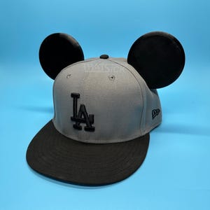 Puede incluir: Gorra de béisbol gris con visera negra y las letras "LA" en negro. La gorra tiene dos grandes orejas circulares negras. Un pequeño logotipo de New Era está en el lateral.