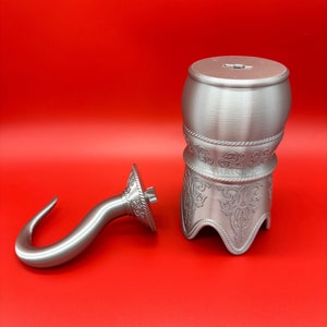 May include: A silver-coloured pirate-themed money box and hook. The money box is barrel-shaped with decorative patterns and a slot on top. The hook is curved with a decorative circular base. Both items are on a red background.