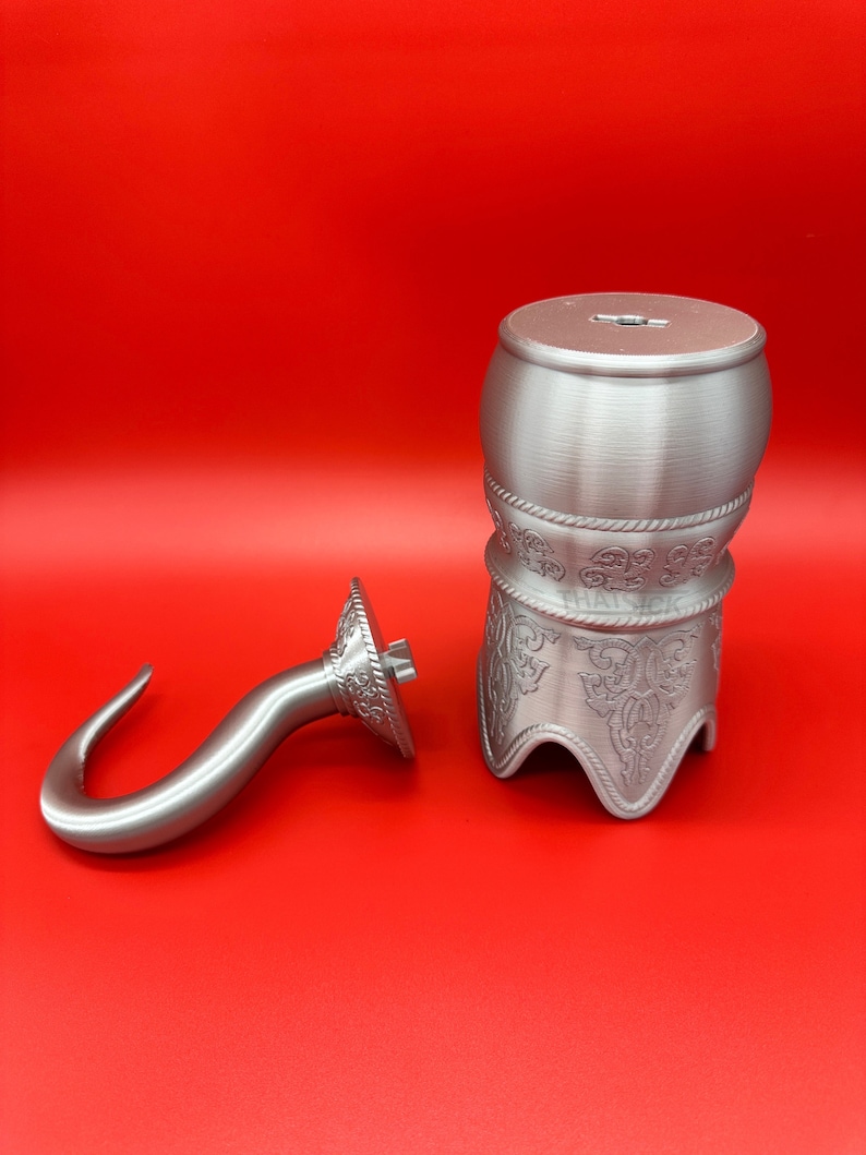 May include: A silver-coloured pirate-themed money box and hook. The money box is barrel-shaped with decorative patterns and a slot on top. The hook is curved with a decorative circular base. Both items are on a red background.