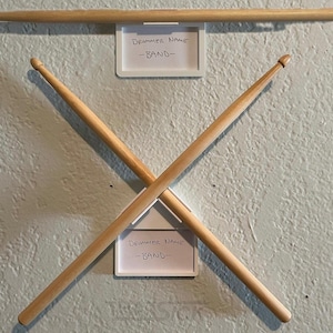 May include: Wooden drumsticks arranged in an X shape, with one drumstick above. Each drumstick is light brown. Two white label holders display the text "Drummer Name" and "-BAND-". The background is a light blue wall.