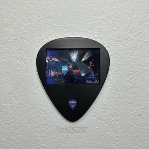 May include: A black guitar pick-shaped frame with a concert photo inside, mounted on a white wall. The photo shows a band performing on stage with bright lights. The text "California Roots" is visible. The text "THAT SICK" is below the frame.