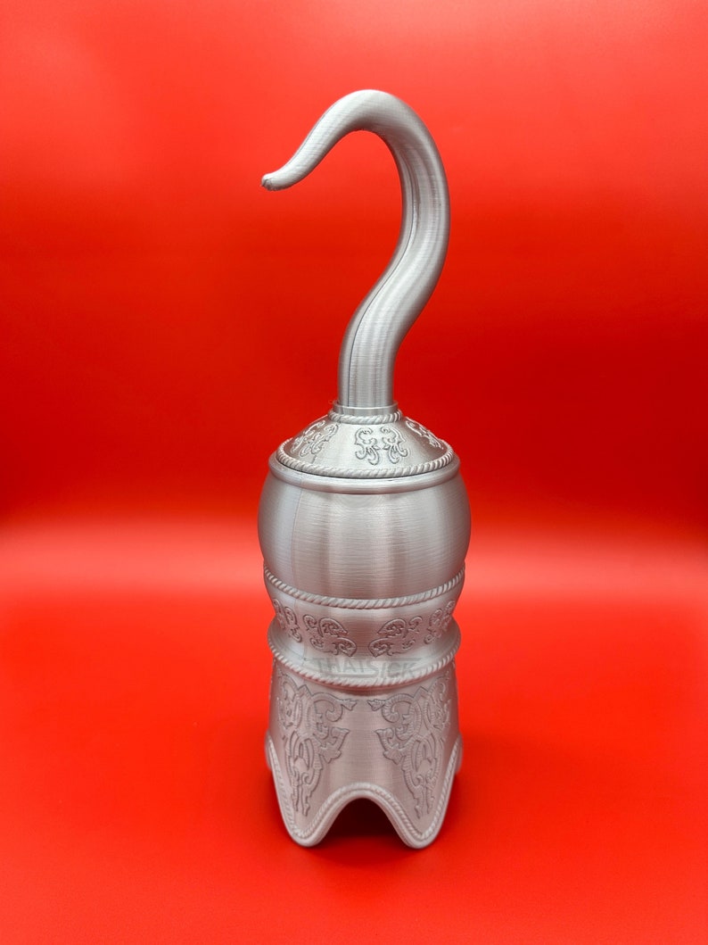 May include: A silver pirate hook with an elaborate design. The curved hook sits atop a base with decorative patterns. The background is a solid red colour.