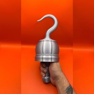 May include: A silver pirate hook prop with a curved hook and a rounded handle. The hook is held against an orange background. The prop is made of metal and has a detailed design.