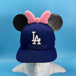 May include: A navy blue baseball cap with the white letters "LA" and the text "THATSICK". The cap is adorned with black mouse ears and a pink bow. The hat is displayed on a white mannequin head.