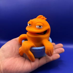 Op de afbeelding: Een oranje, cartoonachtig wezen met een zelfvoldane uitdrukking zit in een kleine, lichtblauwe beker. Het wezen heeft armen en een vin-achtig kenmerk op zijn hoofd. De achtergrond is effen donkerblauw.
