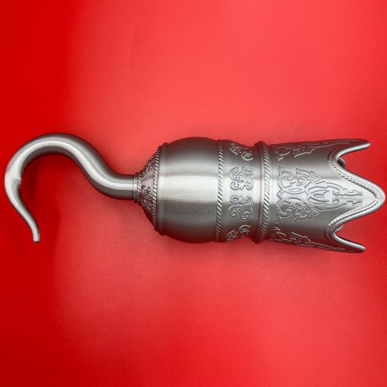 May include: A silver metal hook, possibly a costume prop, set against a red backdrop. The hook features a curved end and a detailed handle with decorative patterns. The handle has a cylindrical form with a flared end.