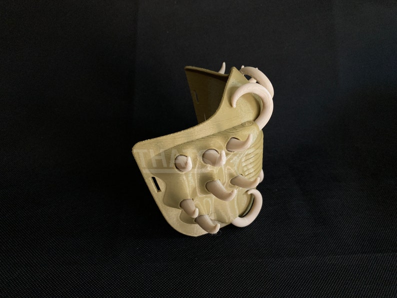 May include: A tan-colored, 3D-printed mask with curved, off-white accents. The mask has a textured surface and a series of protruding, tooth-like shapes. The design suggests a fantasy or cosplay theme.