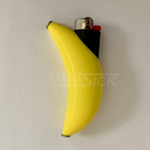 Può includere: Un accendino giallo a forma di banana con base nera e dettagli argentati. L'accendino ha un pulsante di accensione rosso e il testo "THAT SICK" è visibile. L'accendino è posto su uno sfondo bianco.