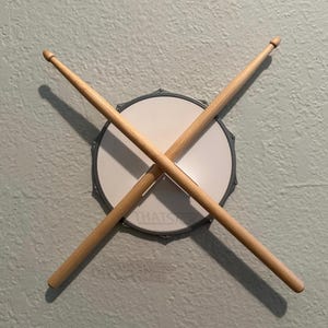 Pode incluir: Uma pele de tambor com baquetas de madeira cruzadas. A pele do tambor é branca com uma borda cinza e o texto "THAT'S IT" impresso. As baquetas são marrons claras e estão posicionadas diagonalmente sobre a pele do tambor.