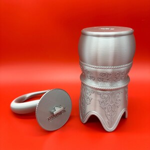 May include: A silver-coloured decorative object with a cylindrical base and a separate, circular piece with a handle. The base features ornate, engraved patterns and the text "THAT SICK" is visible on the circular piece. The background is a solid red.