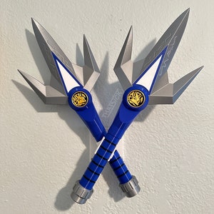 May include: Two crossed blue and silver toy swords. The blades are silver with a white and blue accent. The handles are blue with black stripes and silver accents. Each handle features a gold emblem.